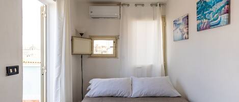 A bedroom with a bed, a window, an air conditioning unit, and two paintings on the wall.