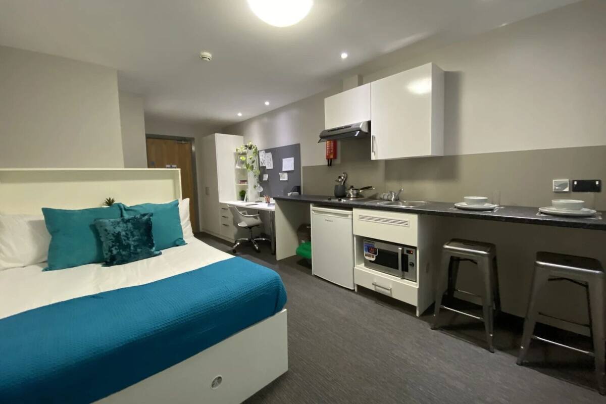 A modern hotel room with a bed, a desk, a kitchenette, and a small bar area.