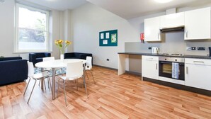 Eine moderne Küche mit weißen Schränken, einem eingebauten Backofen und einer Kaffeemaschine. Ein Essbereich mit rundem Tisch und Stühlen sowie ein Fenster mit Blick auf Grünfläche.