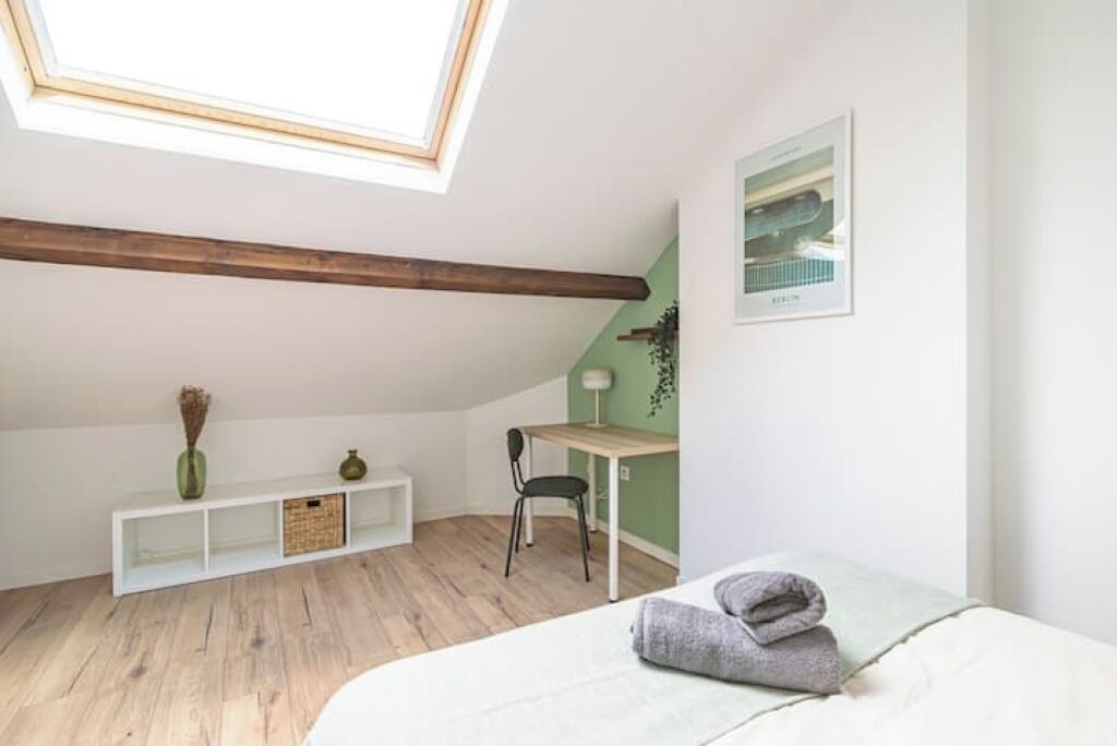 A small, bright room with a sloped ceiling, a desk, a chair, a bed, and a skylight.