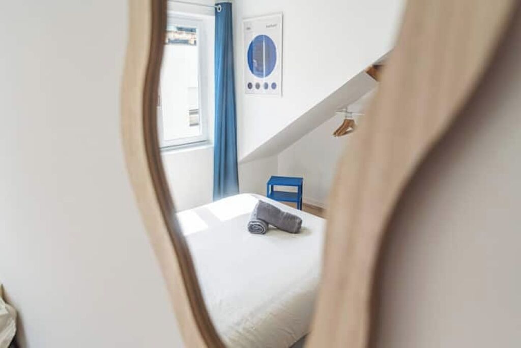 A bedroom with a bed, a window with curtains, a wall-mounted thermostat, and a blue chair.