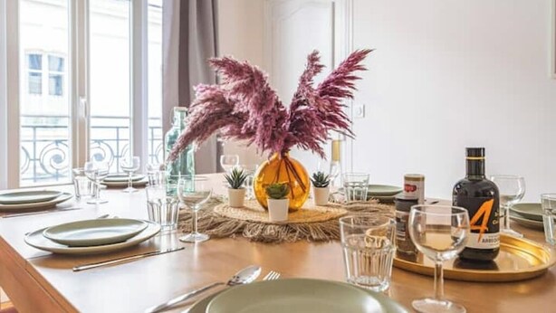 A dining table set with plates, cutlery, and glasses, a bottle of liquor, and a decorative vase with dried plants. - Le Seventies - Exceptional 4 rooms & billiards (Reims)