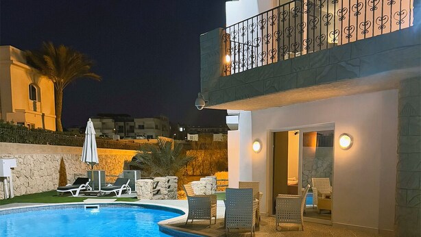 A swimming pool with lounge chairs and umbrellas, a stone wall, and a building with a balcony.