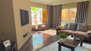 A spacious living room with a sofa, a coffee table, and a TV mounted on the wall. There is a large window with curtains, and a view of a pool and buildings outside.