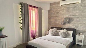 A bedroom with a bed, a nightstand, a wall-mounted air conditioner, and a window with sheer curtains.