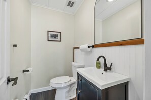 Um banheiro com vaso sanitário branco, pia com espelho acima e quadro pendurado na parede.