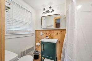 A bathroom with a toilet, a sink, a cabinet, a mirror, and a window with blinds. - The Inn & More (Bartlett)