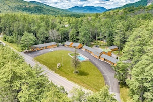 Una cabaña con varias chozas, piscina y estacionamiento rodeado de un bosque denso y montañas.