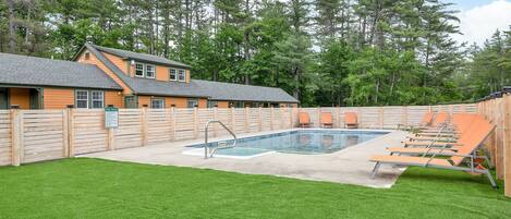 A pool area with a wooden deck, lounge chairs, and a building with a sloped roof.