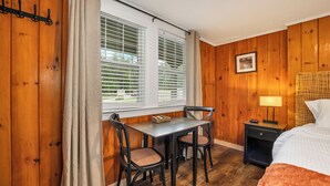 Habitación con paneles de madera, una mesa de comedor y sillas, una cama, una mesita de noche con lámpara y una ventana con persianas.