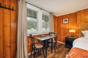 A wooden-paneled room with a dining table and chairs, a bed, a nightstand with a lamp, and a window with blinds. - The Inn & More (Bartlett)