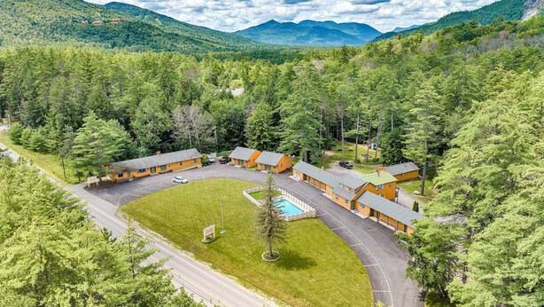Una cabaña con varias chozas, piscina y estacionamiento rodeado de un bosque denso y montañas.