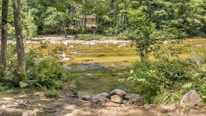 Un río con rocas y helechos, rodeado de árboles y una casa al fondo.