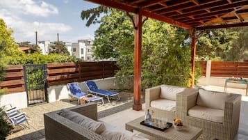 A covered outdoor seating area with wicker furniture, a table with a tray, and a view of buildings and trees in the background.