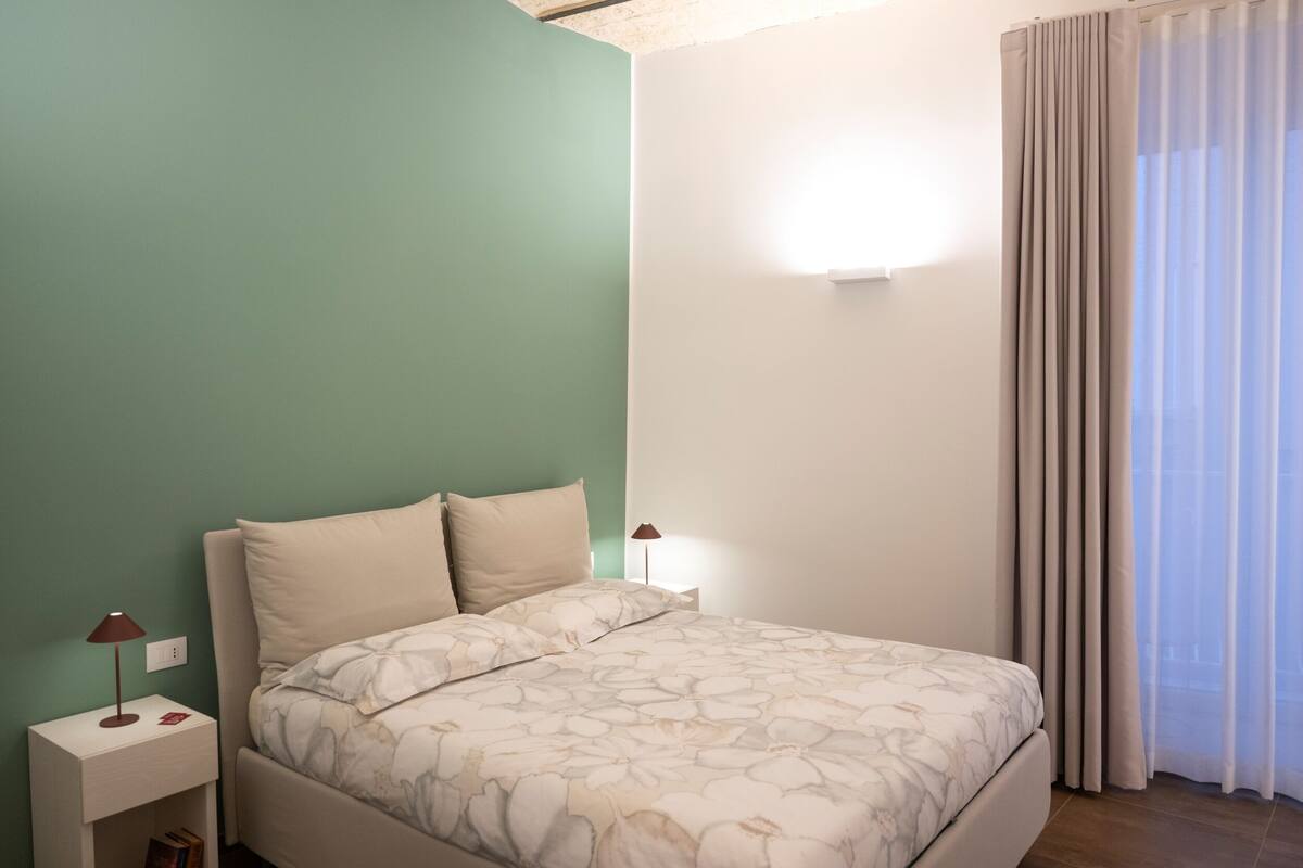 Un dormitorio con cama, mesitas de noche, una lámpara de pared y una ventana con cortinas.