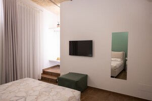 Un dormitorio con cama, un reposapiés verde, televisor montado en la pared y un espejo que refleja la cama.