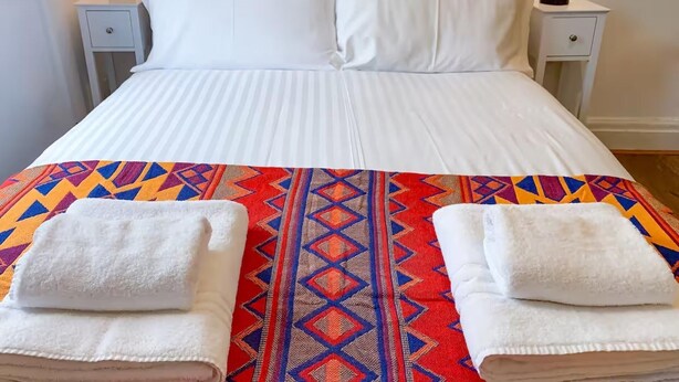 Una cama con sábanas blancas, dos mesitas de noche con lámparas y una manta de colores con estampado geométrico.