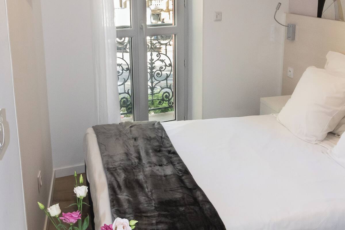 Ein Hotelzimmer mit einem Bett, einem Nachttisch mit Blumenvase, einem an der Wand befestigten Fernseher und einem Fenster mit Blick ins Freie.