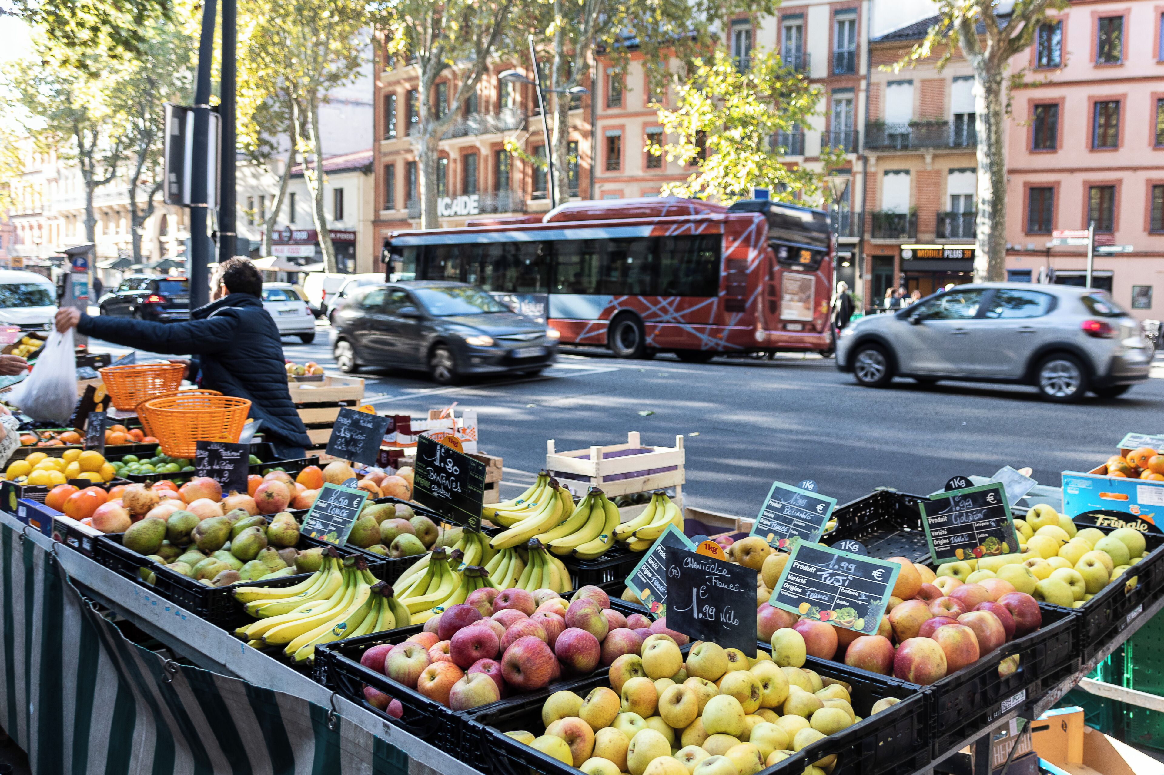 Een straatmarkt met vers fruit, waaronder appels en bananen, en een bus op de achtergrond.