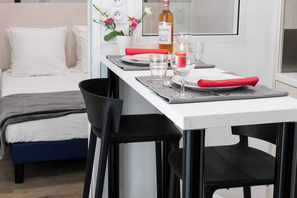 Ein modernes Hotelzimmer mit einem weißen Bett, einem kleinen Esstisch mit Stühlen, einem Spiegel und einer Wand mit abstrakten Kunstwerken.