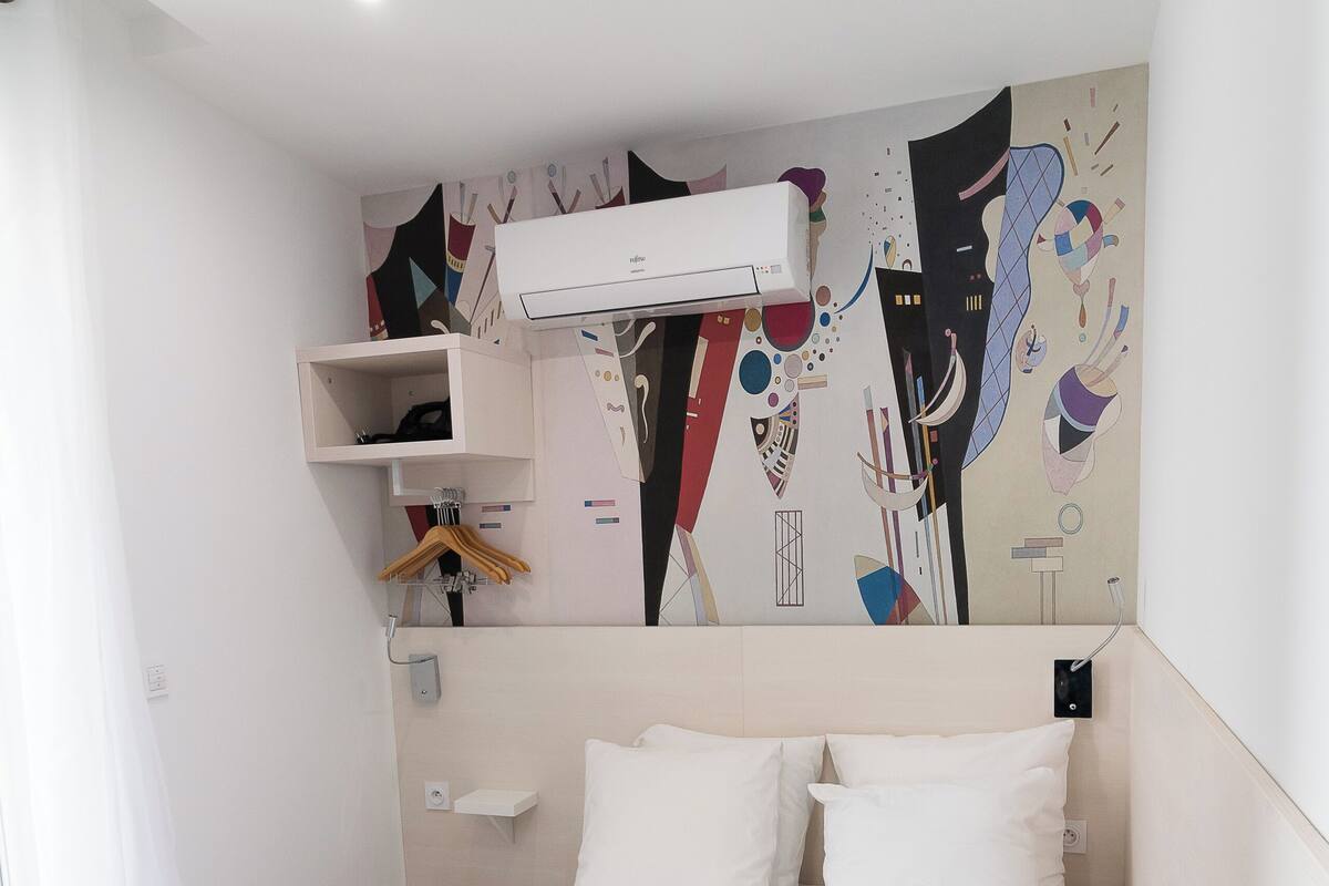 Ein Schlafzimmer mit einem Bett, weißen Kissen, einer grauen Decke, einer Wand-Klimaanlage, einem Regal mit Kleiderbügeln und einem farbenfrohen, abstrakten Wandbild.