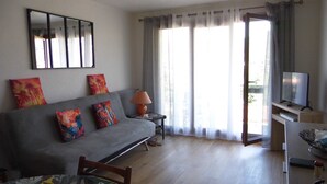 Una sala de estar con un sofá gris, cojines coloridos, una mesita con una lámpara, un televisor sobre un mueble bajo y un ventanal con cortinas translúcidas.