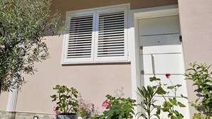 Une maison à l’extérieur de couleur beige, avec une porte blanche et une fenêtre dotée de volets blancs. Des plantes en pot sont disposées devant la maison.