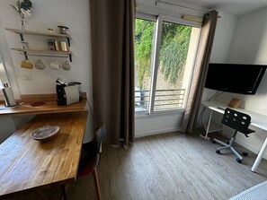 Ein Zimmer mit einem Holztisch, einem Stuhl, einem Fernseher, einem Fenster mit Vorhängen und einem Regal mit Gegenständen.