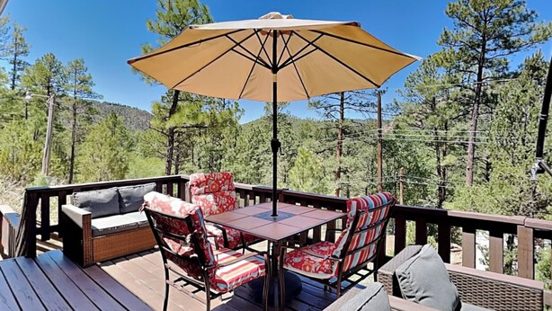 A wooden deck with a table and chairs, a large umbrella, and a view of a forested area. - Knotty Pines Cabin by Ruidoso Vacation Rentals (Ruidoso)
