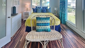 A bedroom with a bed, a chair, a nightstand, a lamp, and a window with curtains. - Jamaica Palms Oasis by Ryson Vacation Rentals (Galveston)