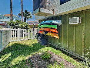 A fenced backyard with a small garden area, a white picket fence, and a row of colorful kayaks hanging on the wall. - Jamaica Palms Oasis by Ryson Vacation Rentals (Galveston)