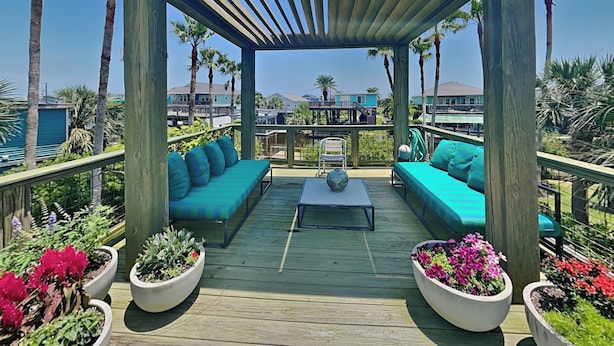 A wooden deck with a covered seating area, surrounded by potted plants and overlooking a residential area. - Jamaica Palms Oasis by Ryson Vacation Rentals (Galveston)