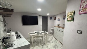 A compact kitchen with a dining area, white marble flooring, a flat-screen TV, and a pizza-themed wall decoration.