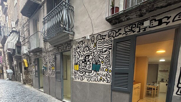 A narrow street with cobblestone pavement, a building with a metal balcony, and a storefront with a colorful mural.