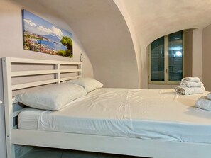 Un dormitorio con cama blanca, un cuadro de un paisaje y una ventana con persianas.