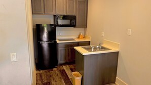 A compact kitchen with wooden cabinets, a black refrigerator, a microwave, a sink, and a trash bin. - Garden Inn Express (Humble)