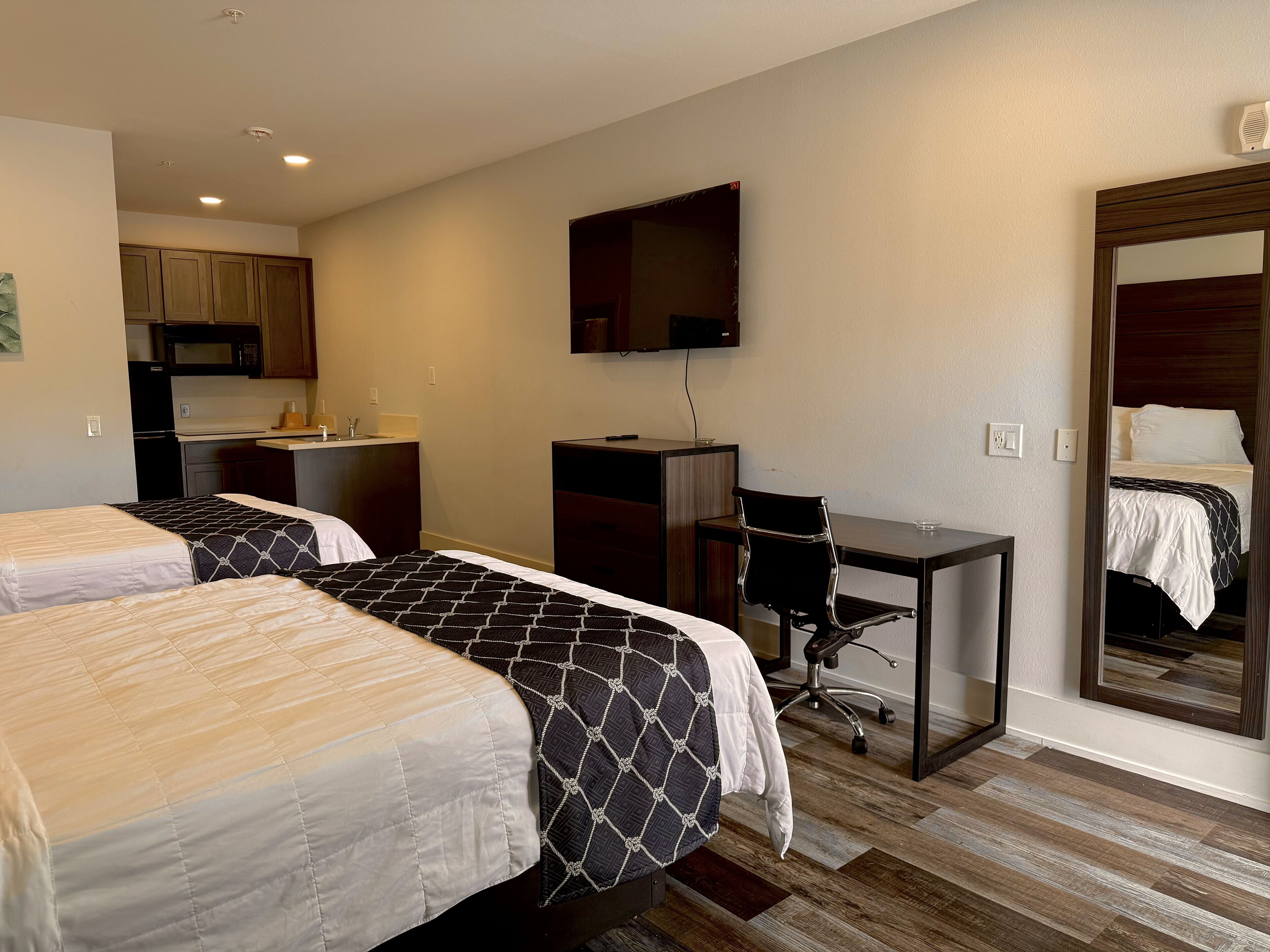 Quarto de hotel com duas camas, uma escrivaninha, uma cadeira, televisão, espelho e cozinha compacta.