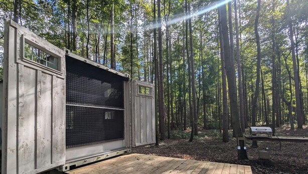 Une cabane rustique avec une terrasse en bois et une porte moustiquaire, entourée d’une forêt.