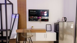 A modern hotel room with a wooden desk, a chair, a refrigerator, and a wall-mounted TV displaying a Netflix show.