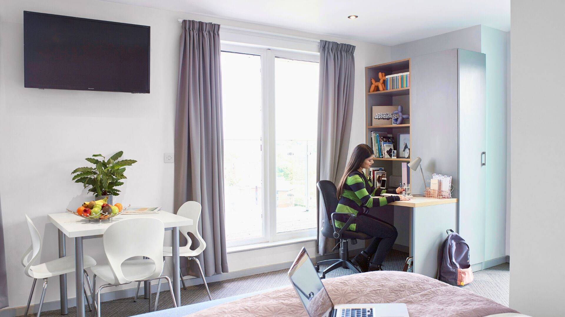 A modern hotel room with a desk, chair, laptop, and a bed.