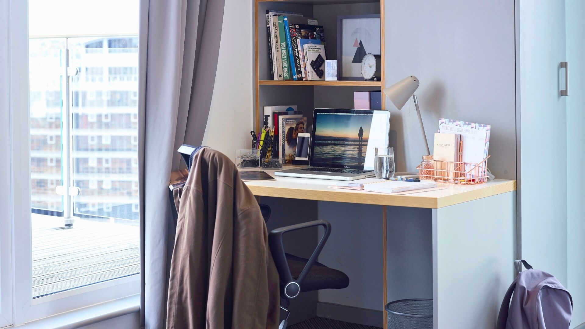 A hotel room with a desk, laptop, books, and a view of buildings through the window.