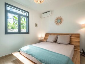Un dormitorio con una cama, una ventana con vistas a la vegetación, aire acondicionado y un adorno en la pared.