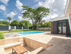 Un área de piscina exterior moderna con piscina azul cristalina, un banco de madera y una escalera que lleva a un espacio de estar cubierto.