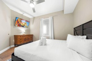 Un dormitorio con una cama, un televisor montado en la pared, un ventilador de techo, una ventana con cortinas y un aparador de madera.
