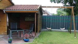 Un cobertizo de madera con techo de tejas, un parque infantil con columpio y una pequeña zona de patio con dos sillas y una mesa.