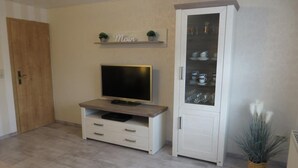 Una sala de estar con un televisor sobre un mueble de madera, un armario blanco con puertas de cristal y un estante pequeño con plantas.