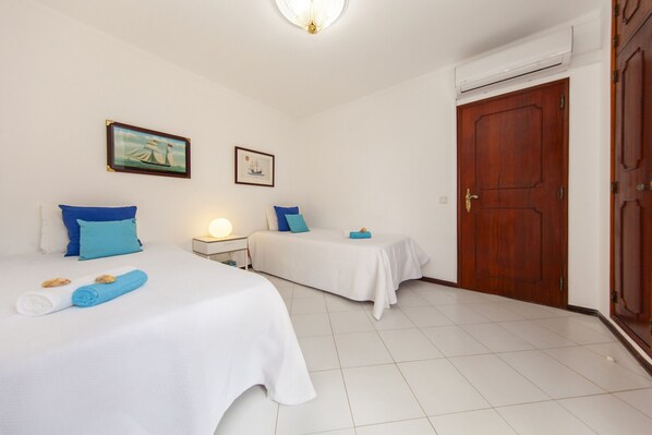 A hotel room with two beds, white bedding, blue pillows, a nightstand with a lamp, and a wooden door.