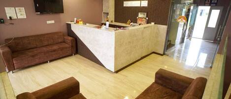 A reception area with a marble counter, brown sofas, a television, and a glass door leading to the toilet.