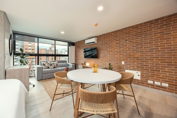 Una sala de estar moderna con una mesa redonda blanca, sillas de mimbre, un sofá gris y un ventanal con vistas a la ciudad.