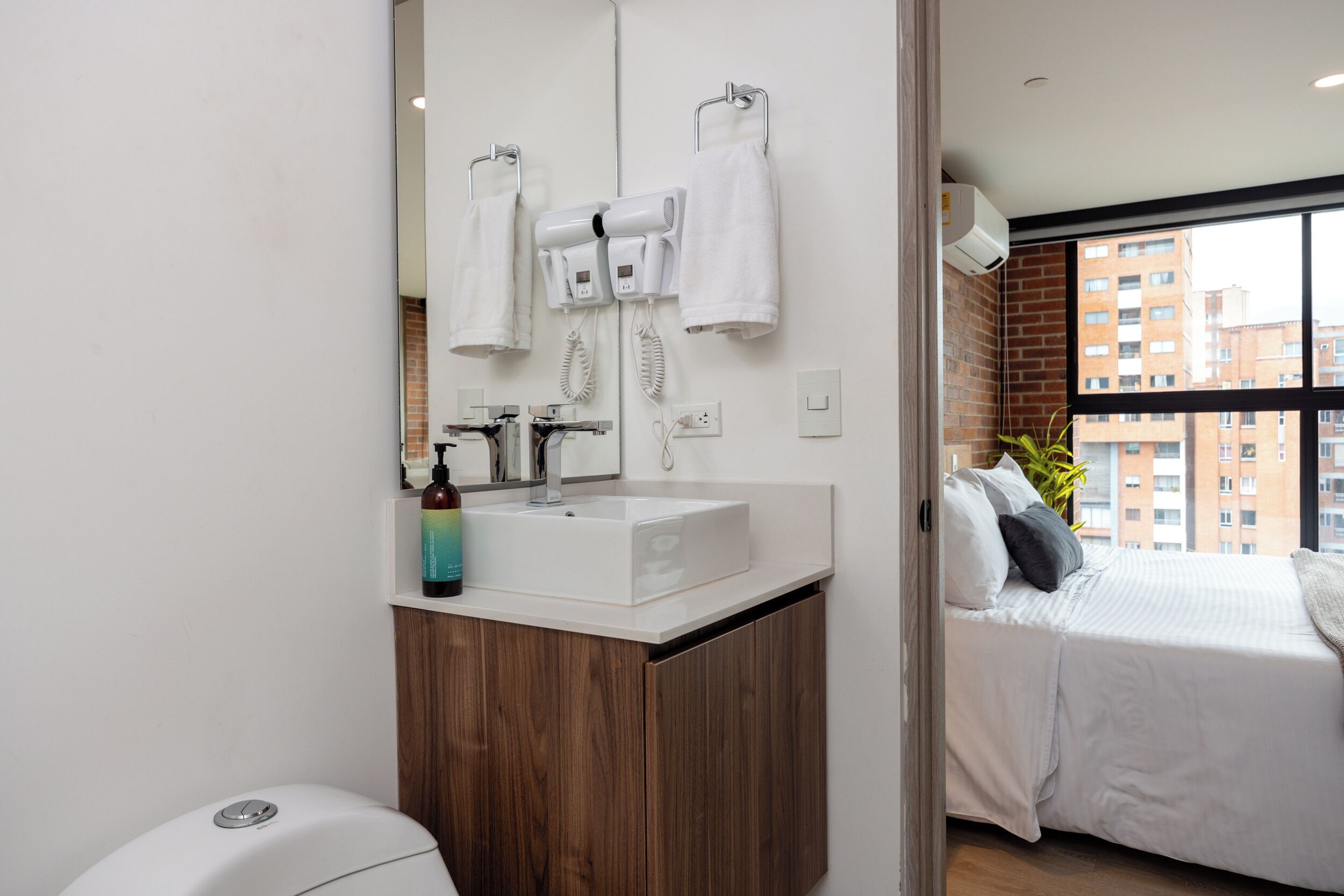 A modern bathroom with a white sink, a mirror, and a hairdryer. There is a bed with a window view of a cityscape.